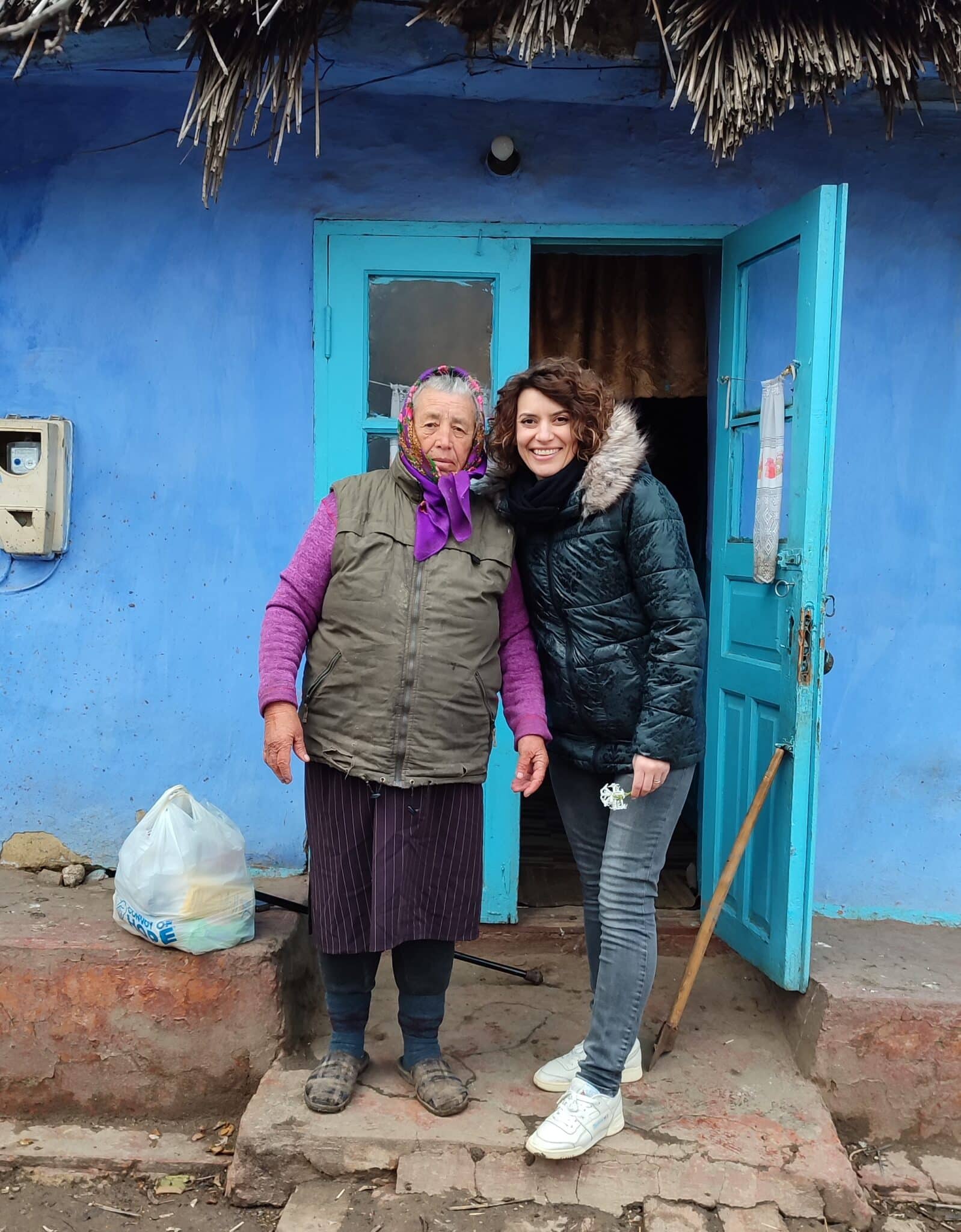 Two women stand in front of the doorway of a house. Both the house and door are painted vibrant hues of blue. There is a bag with 'Convoy of Hope' printed on it on the ground next to one of the women.